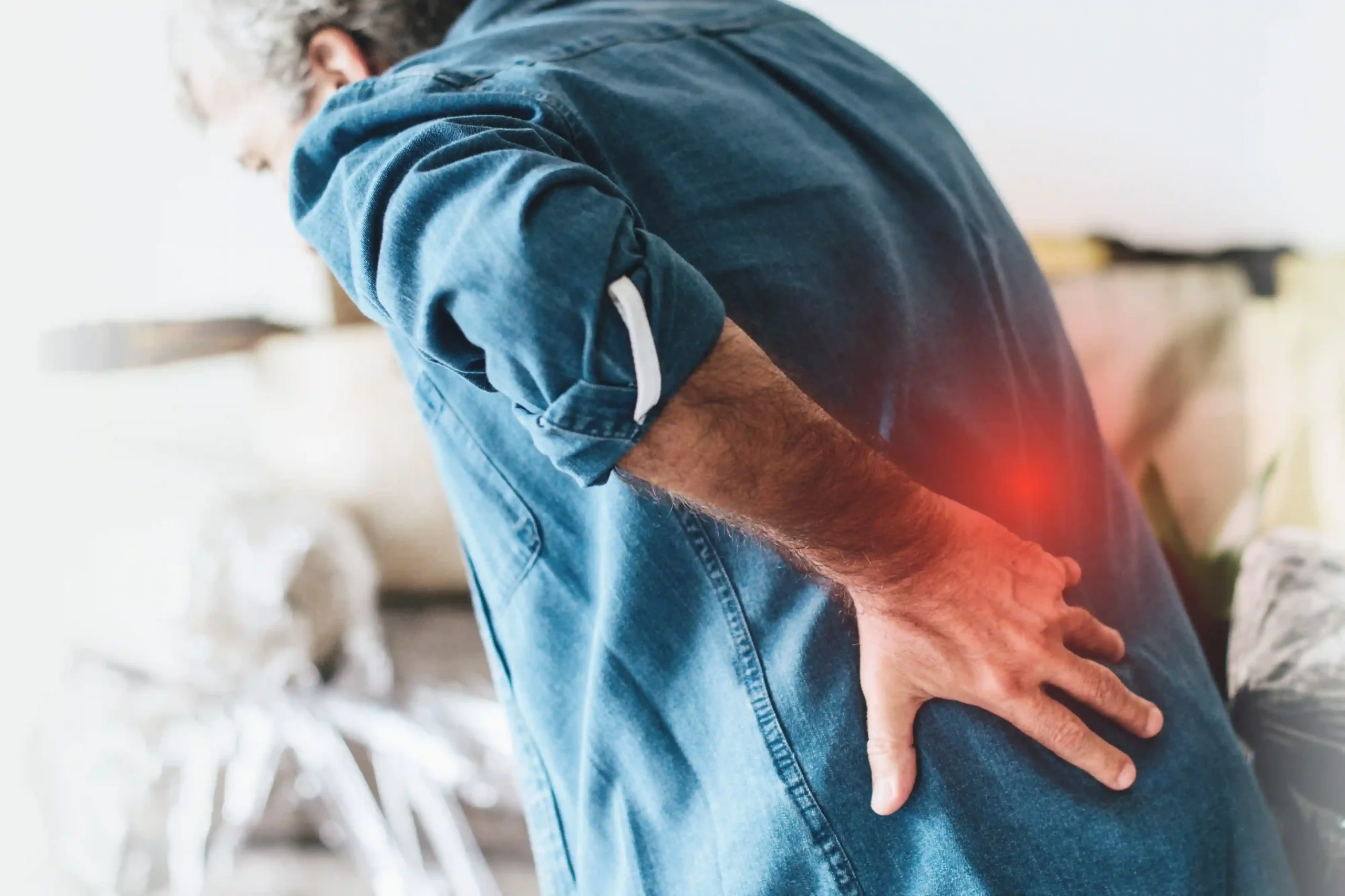 A man in a blue shirt bends forward, pressing his hand against his lower back as a red glow highlights pain, suggesting discomfort or injury.