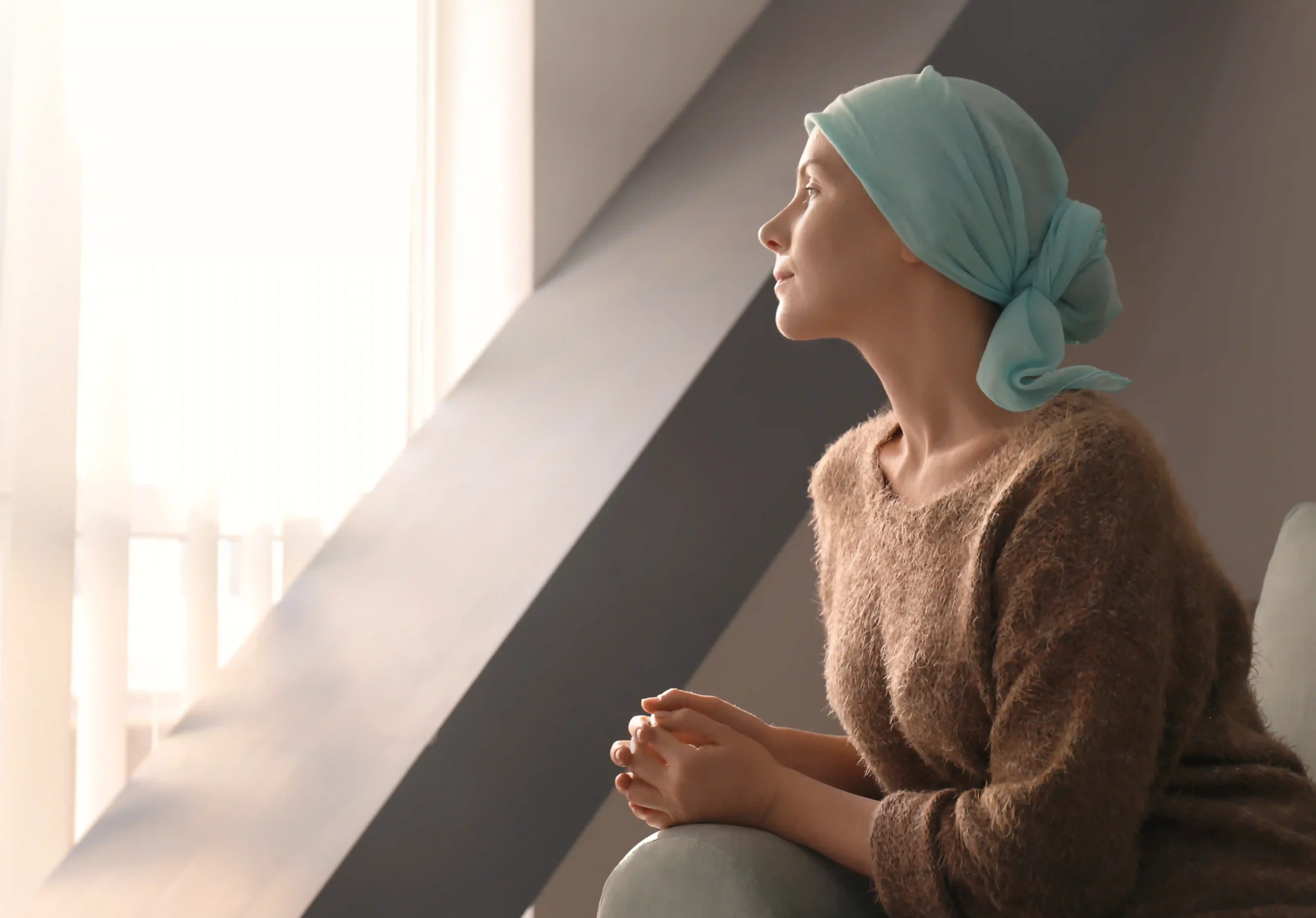 Woman wearing a headscarf sitting by a window while undergoing cancer treatment.