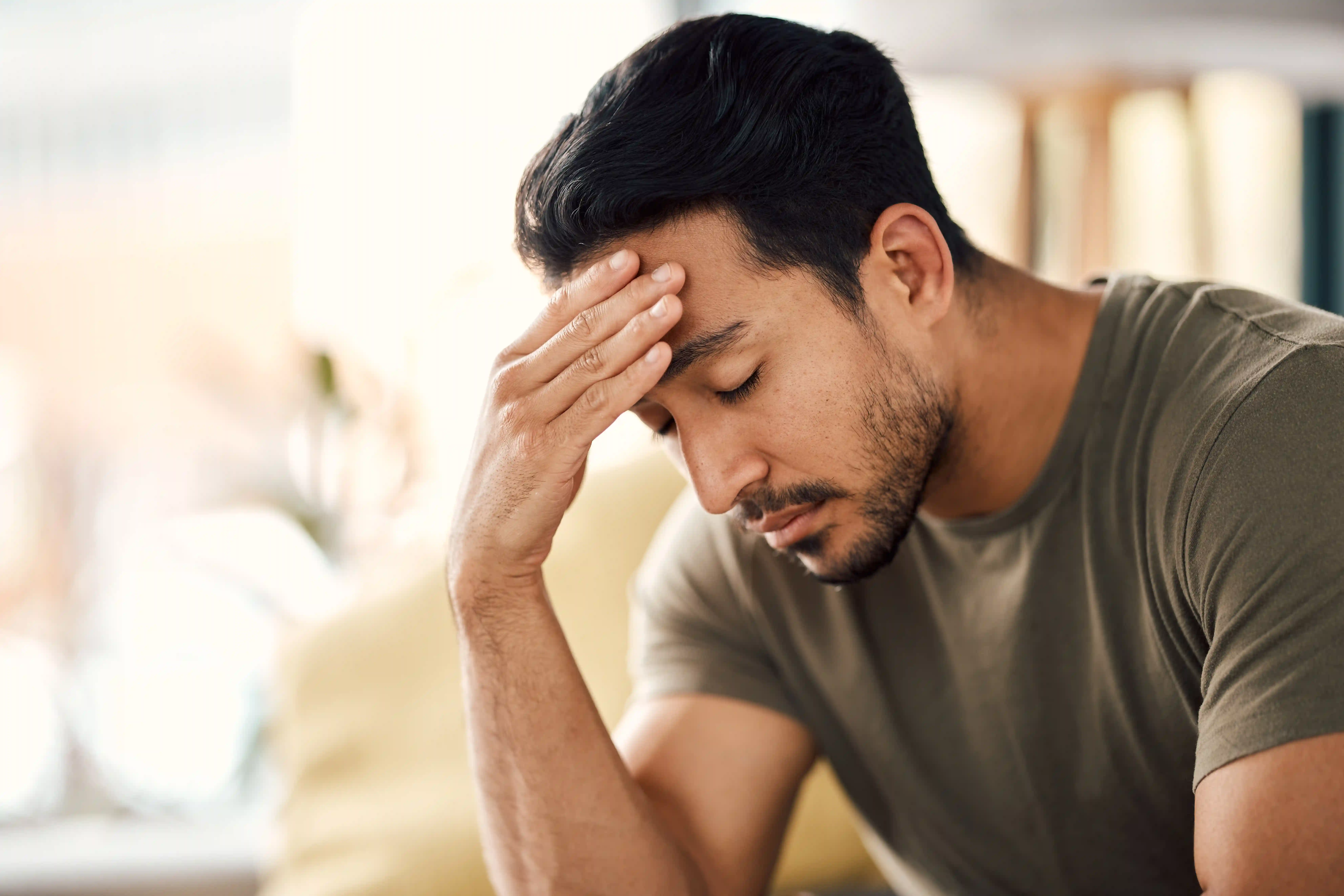 A young man sits indoors, eyes closed and hand pressed to his forehead, appearing stressed or exhausted in a softly lit home setting.