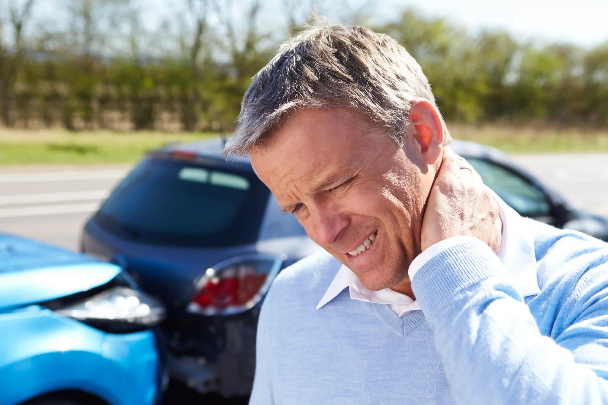 Man experiencing neck pain after a motor vehicle accident with damaged cars in the background.